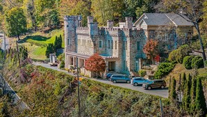 Un edificio de estilo castillo con muros de piedra y múltiples torres, rodeado de exuberante vegetación y árboles.