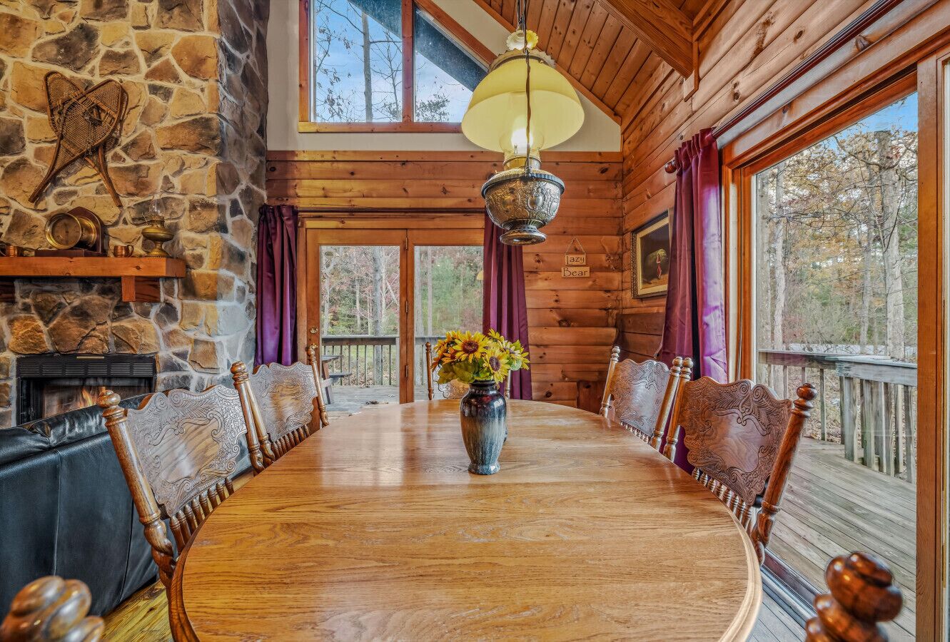 Una mesa de comedor de madera con sillas, un jarrón con flores, una chimenea de piedra y un ventanal con cortinas.