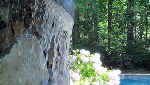 A stone water feature with cascading water, a pool, and pink flowers in a garden setting.