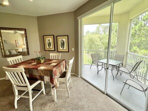 A dining area with a table covered by a patterned tablecloth, surrounded by white chairs. There are framed botanical prints on the wall, and a large window provides a view of greenery outside. - 19-202 Legacy Dunes, minutes from Disney! Senior/Family/Kid Friendly! Relax (Kissimmee)