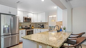 A modern kitchen with white cabinets, stainless steel appliances, a granite countertop, and wicker chairs. - Gorgeous Vacation Dream Home with heated Pool (Seminole)
