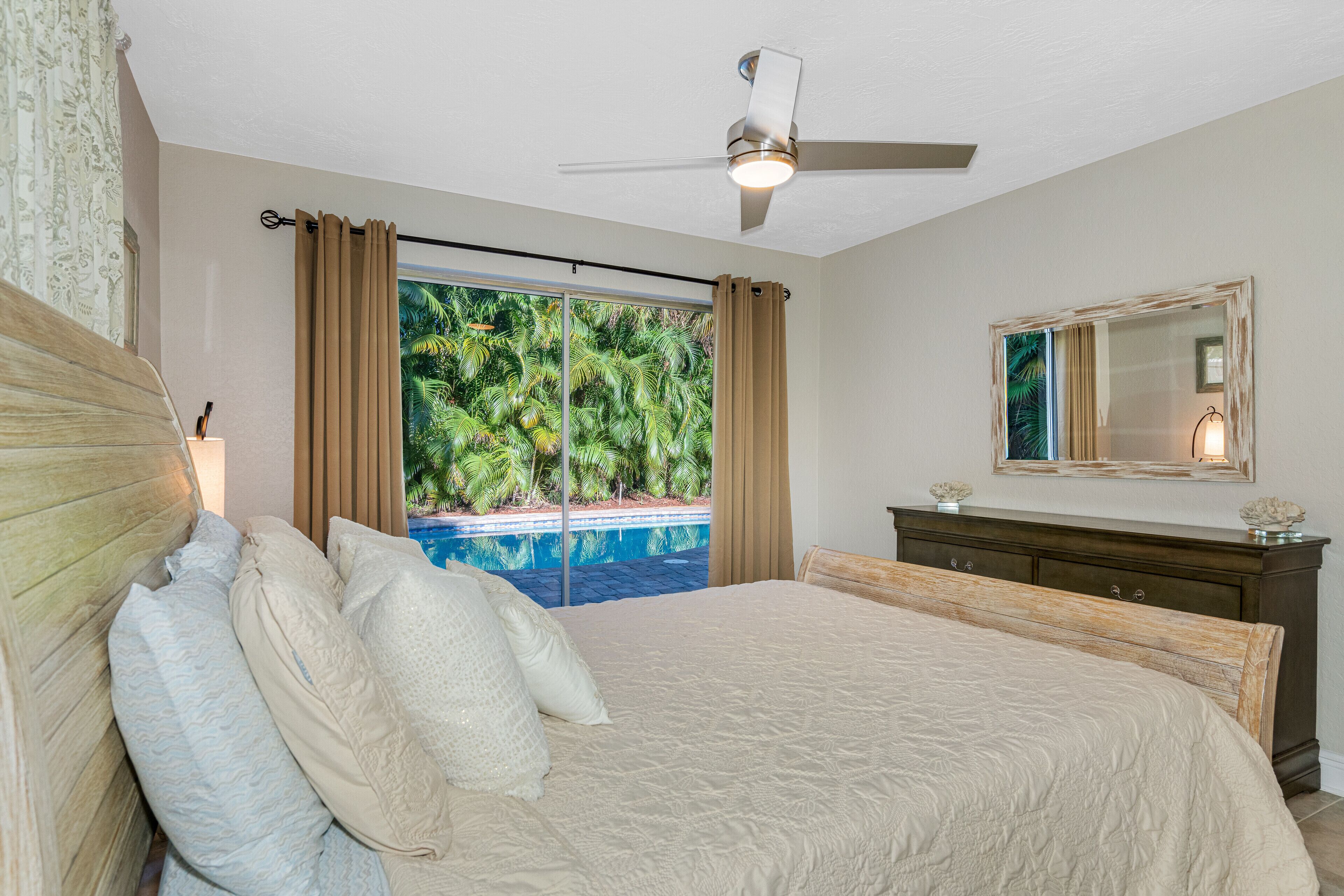 A bedroom with a bed, a ceiling fan, a view of a swimming pool, and a dresser with a mirror.