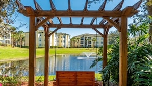 Ein hölzernes Schaukelgestell unter einer Pergola mit Blick auf einen Teich und Gebäude im Hintergrund.