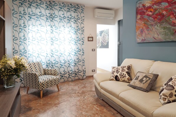 Una sala de estar con cortinas estampadas, un sofá color beige con cojines decorativos, un sillón con diseño y un jarrón con flores.