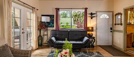 A living room with a black leather sofa, a coffee table with flowers, a large window with curtains, and a door leading to a garage.