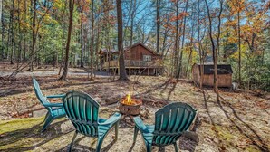 A cabin with a deck, surrounded by a forest with autumn foliage. There are two chairs facing a campfire.