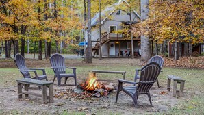 Un espace extérieur rustique aménagé avec des chaises Adirondack et un foyer, entouré de feuillages d’automne et avec un chalet en arrière-plan.
