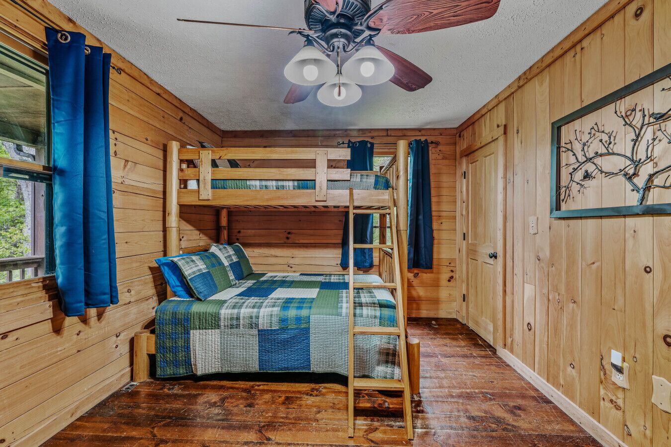 Une chambre avec des lits superposés, un ventilateur de plafond en bois, une couvre-lit matelassée et une vue sur l’extérieur par une fenêtre.