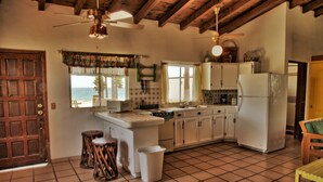 A kitchen with a tiled floor, wooden ceiling beams, a refrigerator, a stove, and a window overlooking the ocean.