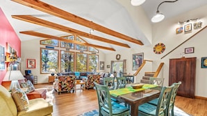 A spacious living room with a large window, wooden beams, a dining area with a glass table, and a staircase. - Healing House - fireplace, wifi, 2 kings, furnished den, screen porch, pool access (Berkeley Springs)