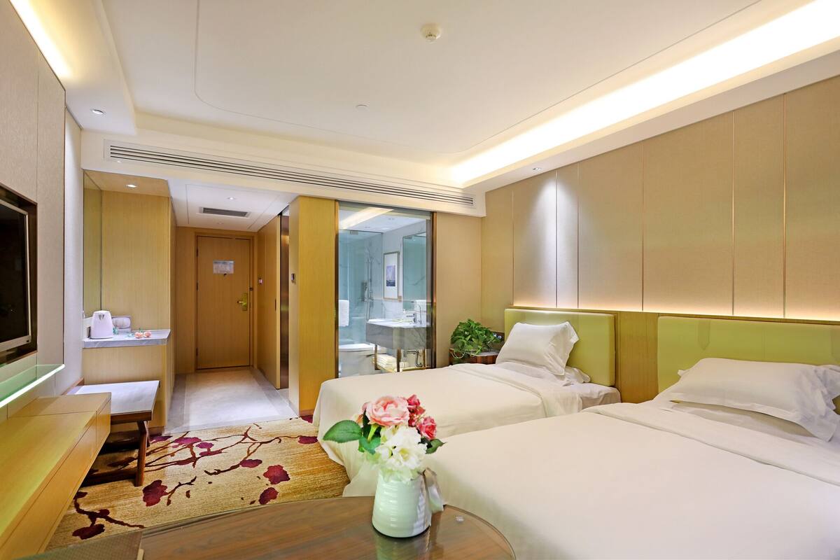 Habitación de hotel con dos camas, televisor, escritorio y baño visible a través de una puerta de cristal.