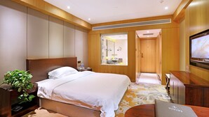 A hotel room with a large bed, a wooden headboard, a bedside table with a plant, a TV, and a bathroom visible through a glass door. - Yantai Asia Hotel (Yantai)