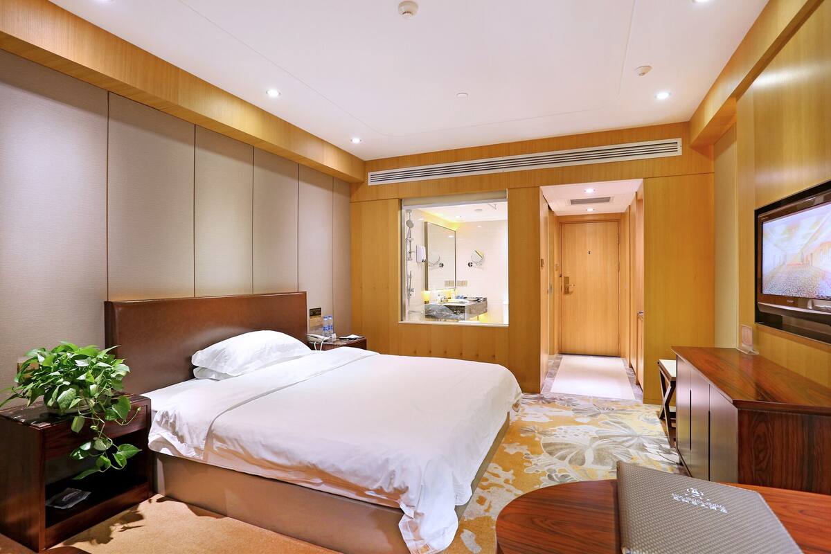 Habitación de hotel con una cama grande, cabecera de madera, mesita de noche con una planta, televisor y baño visible a través de una puerta de cristal.