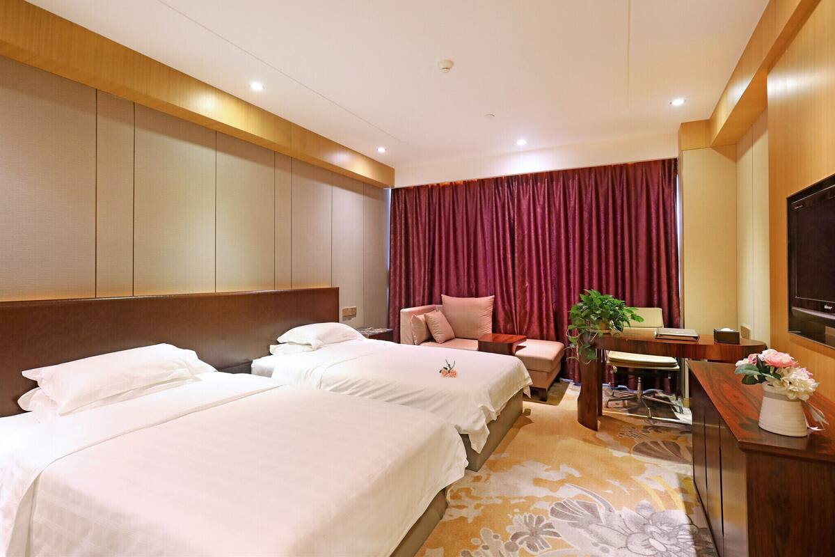 Habitación de hotel con dos camas, un escritorio, una silla, un televisor y un ventanal grande con cortinas.