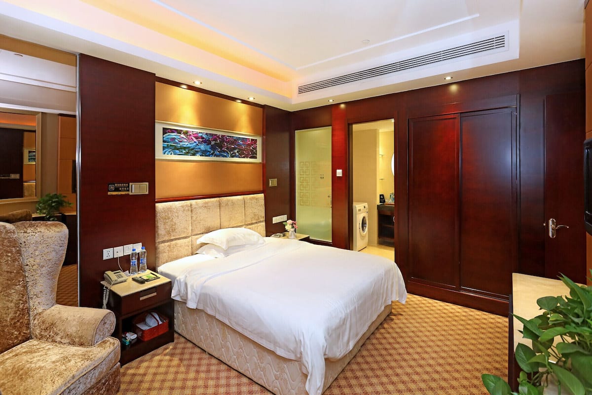 Habitación de hotel con una cama grande, una mesita de noche con un teléfono y botellas de agua, una silla, un cuadro en la pared y un armario.