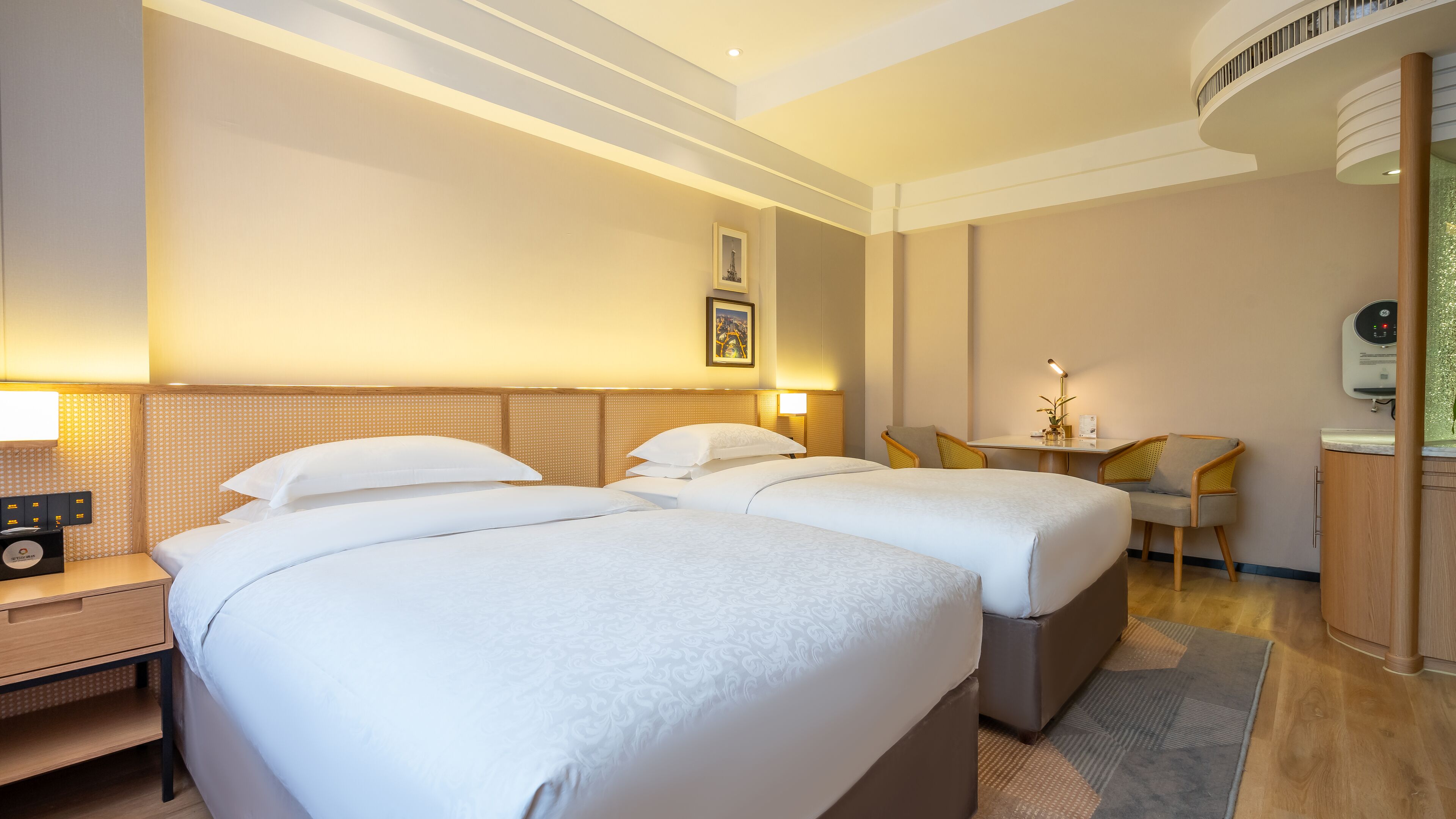 A hotel room with two beds, a desk, a chair, and a small table with a lamp.
