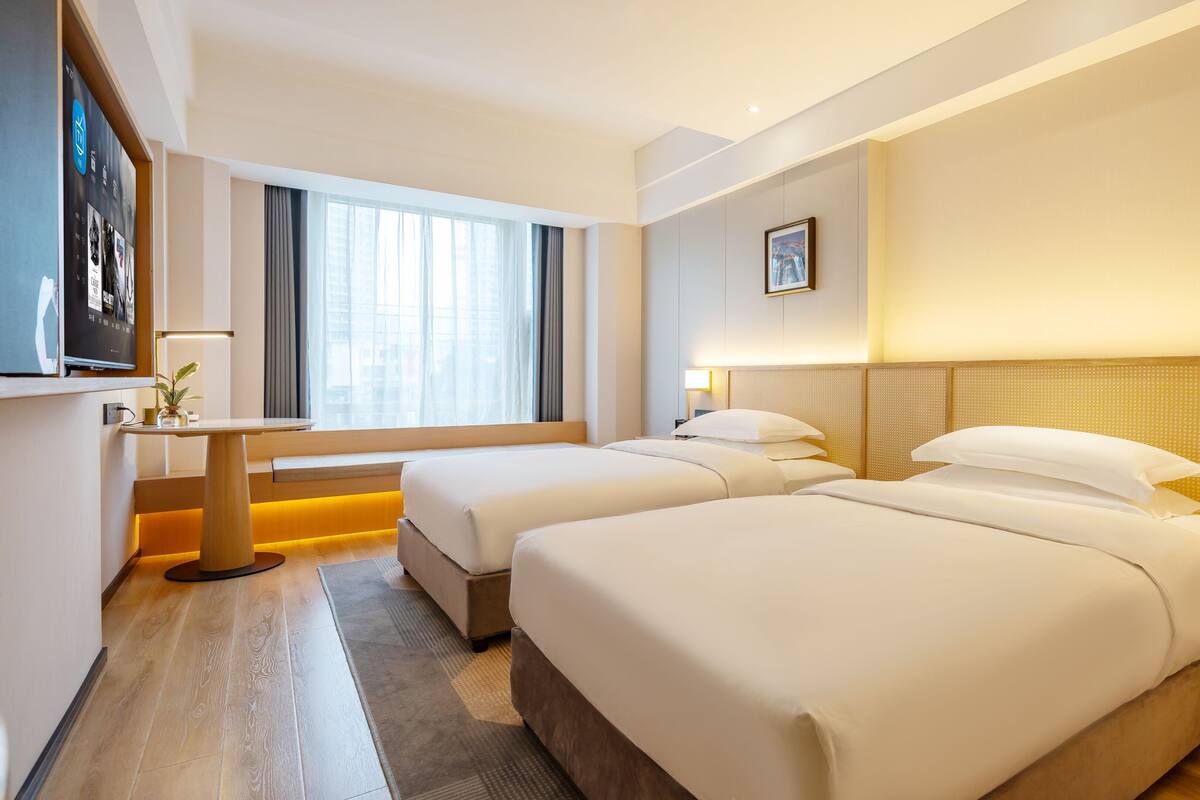 Ein Hotelzimmer mit zwei Betten, einem Schreibtisch, einem Fernseher und einem Fenster mit Vorhängen.