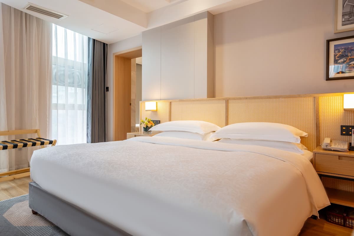 Ein Hotelzimmer mit einem großen Bett, einem Nachttisch mit Lampe, einem Holznachtkästchen und einem Fenster mit durchscheinenden Vorhängen.