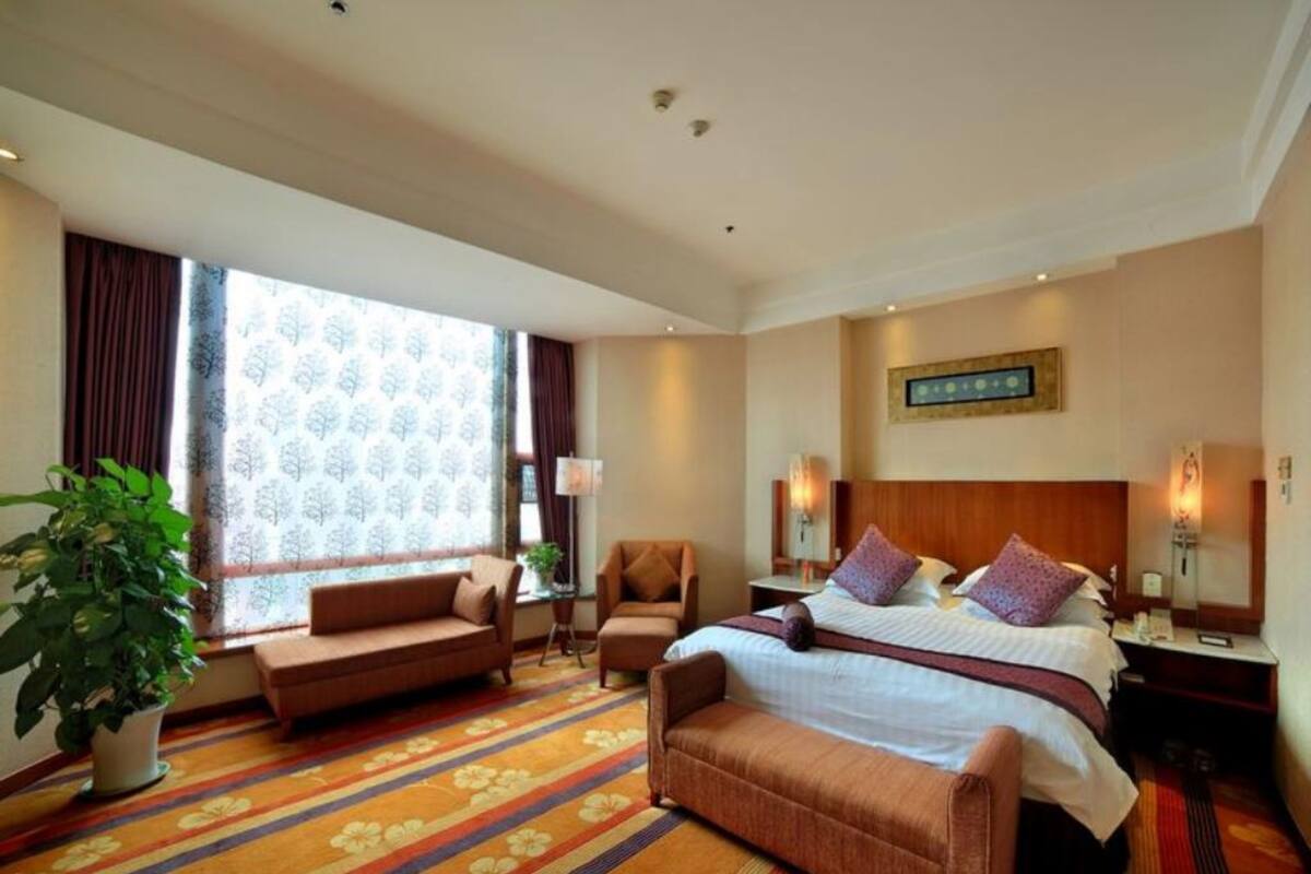Ein Hotelzimmer mit einem großen Bett, zwei Sesseln, einem Sofa, einer Topfpflanze und einem großen Fenster mit Vorhängen.
