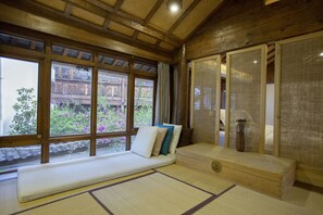 A traditional Japanese room with tatami flooring, a low wooden bed with cushions, a sliding door, and a view of a garden through large windows.