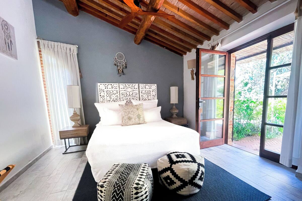 A bedroom with a large bed, two ottomans, a nightstand, a lamp, and a view of greenery through a glass door.