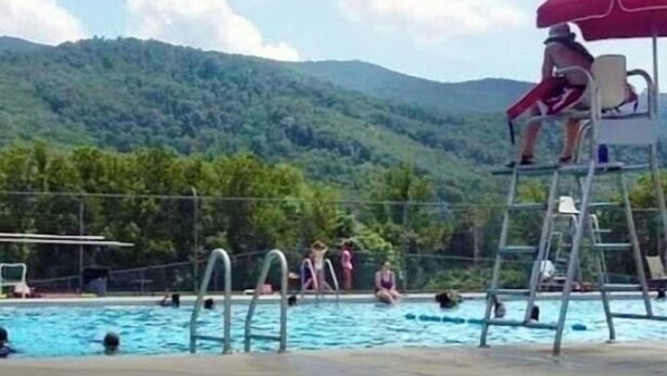 A swimming pool with a lifeguard chair and a red umbrella, surrounded by a forested hillside.