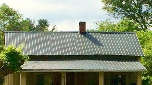 A rustic cabin with a metal roof and a porch. There are four Adirondack chairs arranged around a small fire pit.