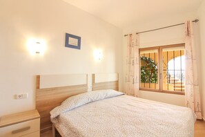 Quarto com cama, mesa de cabeceira, luz na parede, janela com cortinas e vista para o verde lá fora.