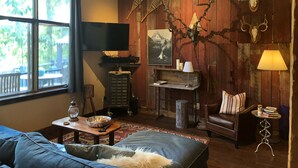 A cozy living room with a large window, wooden walls, a television, a fireplace, and a leather sofa. - The Wild Mile River Retreat (Bigfork)