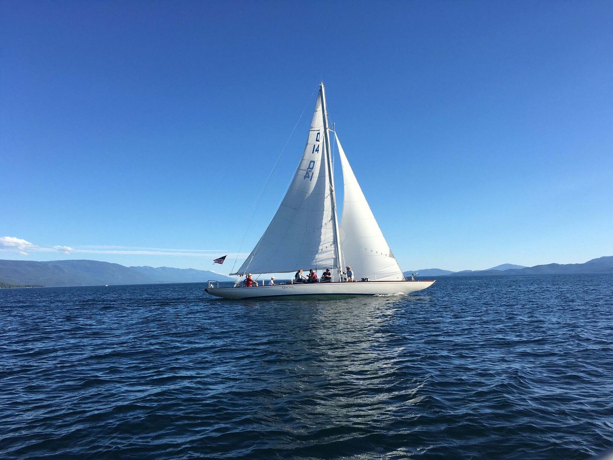 Un voilier à la coque et aux voiles blanches, naviguant sur une étendue d’eau calme, avec des montagnes lointaines se découpant sur un ciel bleu clair.