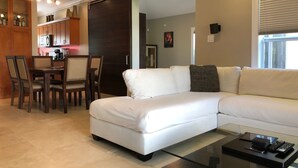 A modern living room with a white sectional sofa, a glass coffee table, and a dining area with wooden chairs and a kitchen in the background.