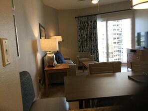 A living room with a dining table, sofa, and armchairs. There is a lamp, a television, and a window with curtains.