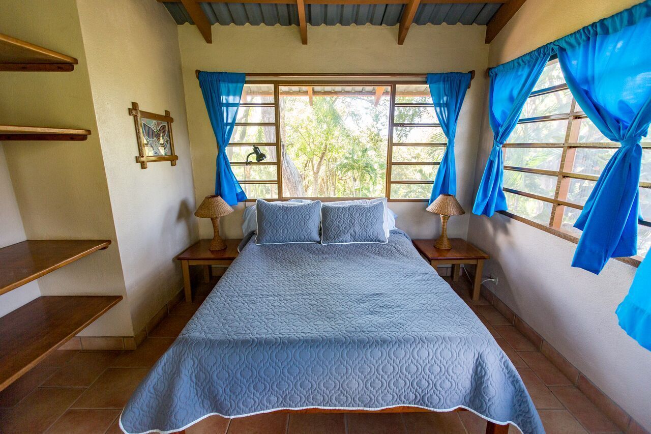 Een slaapkamer met een bed, twee nachtkastjes, een raam met gordijnen, een foto aan de muur en een houten plank.