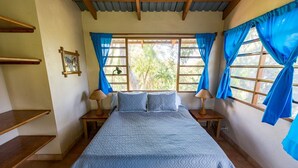 Une chambre à coucher avec un lit, deux tables de chevet, une fenêtre avec des rideaux, un tableau au mur et une étagère en bois.