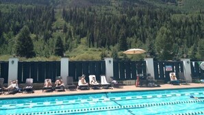 En swimmingpool med liggestole og parasoller, med et skovklædt bjerglandskab i baggrunden.