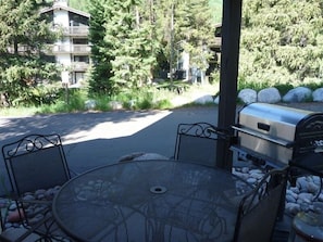 An outdoor patio with a round table and chairs, a grill, and a view of a building and trees.