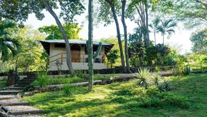 A small, single-story building with a tiled roof, surrounded by lush greenery and a stone pathway leading to it.