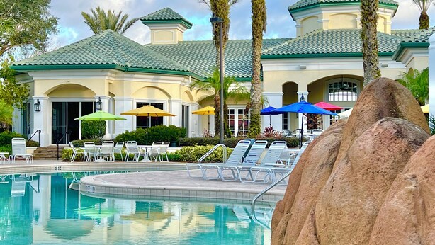 A pool area with lounge chairs and umbrellas, surrounded by palm trees and a building with a green roof. - 1-103 Legacy Dunes, Steps to Pool, Minutes to Disney, King Bed (Kissimmee)