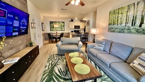 A modern living room with a flat-screen TV, grey sofas, a wooden coffee table, and a dining area with chairs and a table. - 1-103 Legacy Dunes, Steps to Pool, Minutes to Disney, King Bed (Kissimmee)