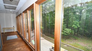 A room with large windows, wooden flooring, a small table, and a view of a forest.