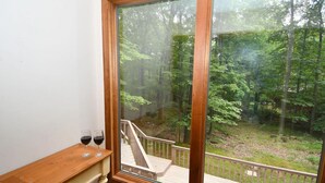 A room with a large window overlooking a forested area. There is a wooden bench with two glasses of red wine on it.