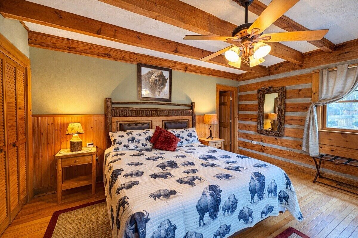 Een slaapkamer met een houten plafond, een bed met een sprei met een buffalobehang, een plafondventilator en een raam met gordijnen.