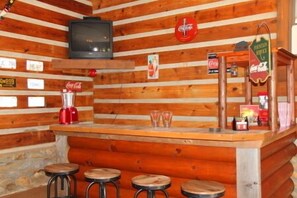 A rustic cabin interior with a wooden bar, stools, a television, and various signs including Coca-Cola branding. - Yes, There Are Mini Donkeys! Peaceful Mountain Cabin Retreat (Clyde)