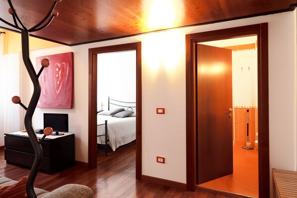 A bedroom with a bed, bedside table, and a painting. There is a door leading to another room.