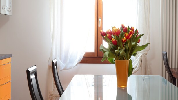 Ein Essbereich mit einem Glastisch, Stühlen, einer Blumenvase und einem Fenster mit Vorhängen.