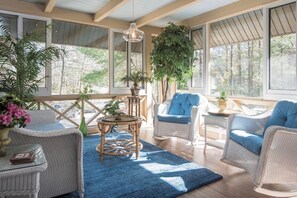 A sunlit room with wicker furniture, a blue rug, and large windows offering a view of trees.