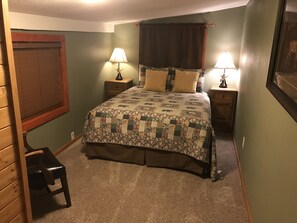 Une chambre à coucher comprenant un lit, deux tables de chevet avec des lampes, une chaise, une fenêtre avec des stores et une porte.