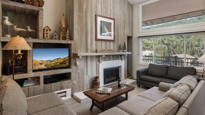 A living room with a fireplace, a television, and a large window overlooking a forest. - Vantage Point 501 - 3 Bedroom Gold (Vail)