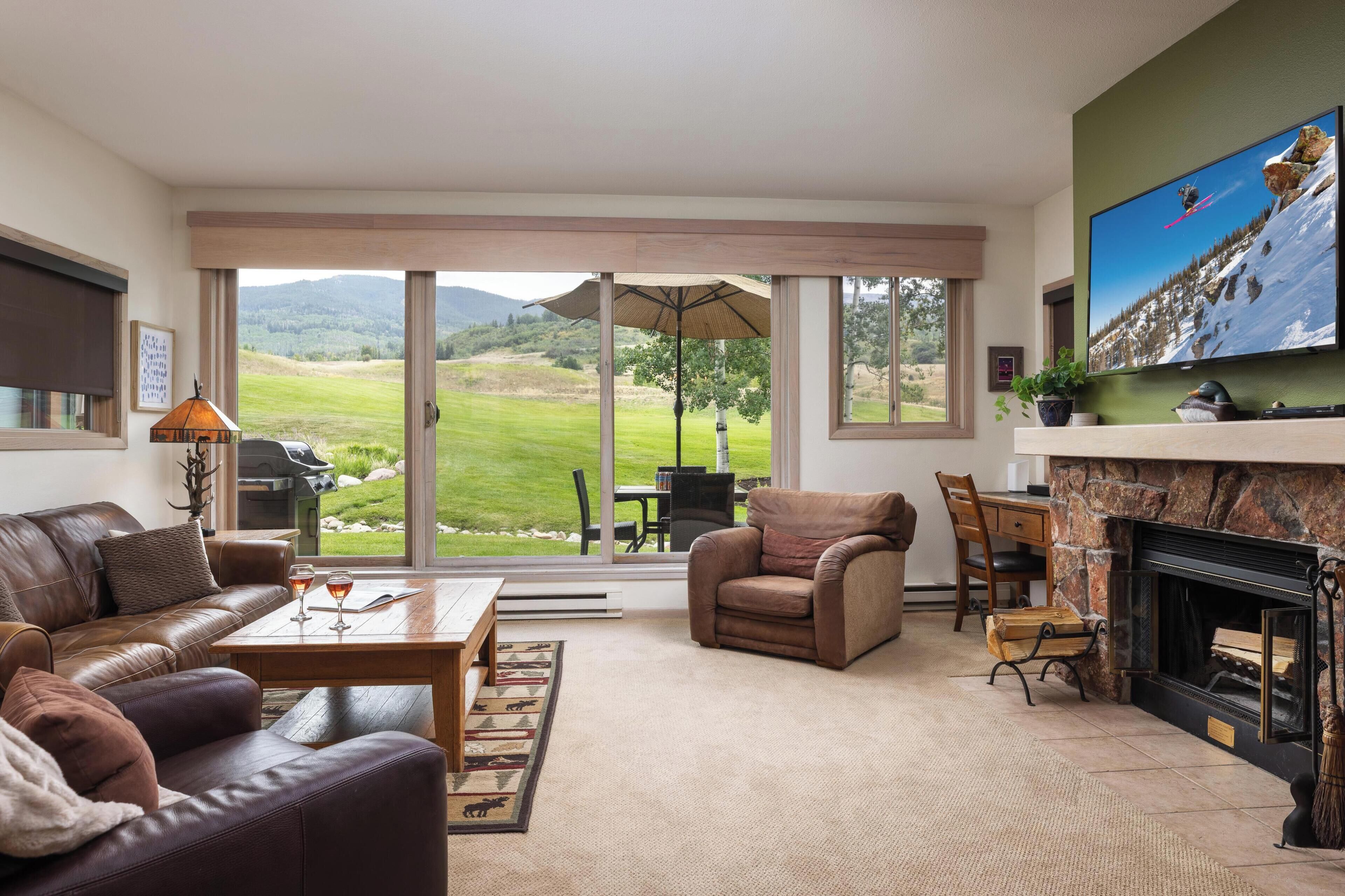 A spacious living room with a leather sofa, a fireplace, a television, and a large window offering a view of a green landscape.