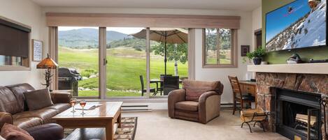 A spacious living room with a leather sofa, a fireplace, a television, and a large window offering a view of a green landscape.